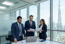 Three professionals in navy suits review documents around a laptop in a glass-walled conference room with a city skyline outside.