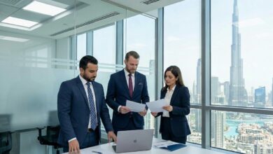 Three professionals in navy suits review documents around a laptop in a glass-walled conference room with a city skyline outside.