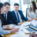 Team of professionals reviewing financial charts around a laptop with bar and line graphs in a bright conference room