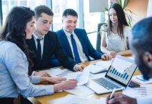 Team of professionals reviewing financial charts around a laptop with bar and line graphs in a bright conference room